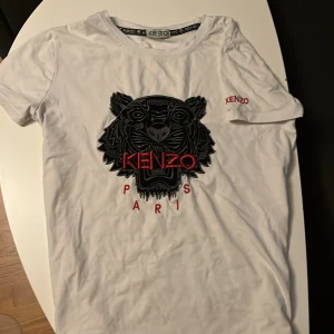 Vit Kenzo t-shirt med tigerbrodyr - Vit t-shirt från Kenzo med ikonisk svart tigerbrodyr och röd broderad logga på bröstet och ärmen. Klassisk passform och korta ärmar, tillverkad i mjuk bomull. Perfekt statement-plagg för dig som vill sticka ut.