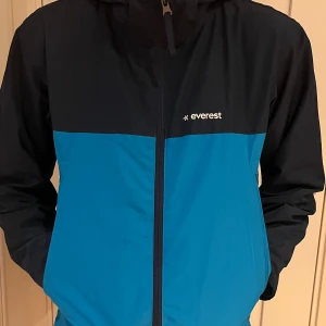 Blå svart regnjacka Everest - Snygg tvåfärgad regnjacka från Everest i blått och svart. Jackan har huva, hel dragkedja framtill och sidfickor. Tillverkad i 100% polyester med foder i nylon. Perfekt för blåsiga dagar och outdoor-aktiviteter. Mycket fint skick