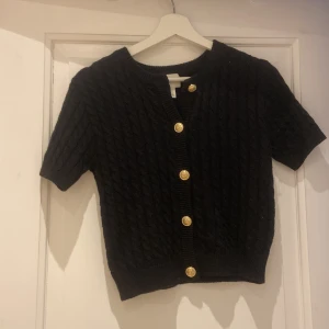Svart kabelstickad kofta från H&M - Svart kabelstickad kofta från H&M med korta ärmar och snygga guldknappar framtill. Koftan har rund halsringning och är i en mjuk stickad kvalitet. Perfekt att slänga över en topp för en chill men ändå uppklädd look.