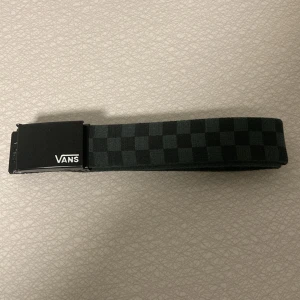 Svart rutigt bälte från Vans - Snyggt svart bälte från Vans med klassiskt rutigt mönster och metallspänne med Vans-logga. Bältet är gjort i slitstarkt tygmaterial och har en stilren, streetinspirerad look som passar perfekt till jeans eller shorts.