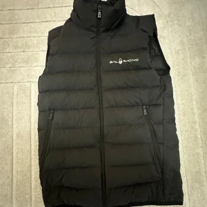 Svart dunväst från Sail Racing - Säljer en svart dunväst från Sail Racing med hög krage och dragkedja framtill. Västen har två fickor med dragkedja och logga på bröstet. Fylld med 800 fill power dun från Allied Feather + Down. Perfekt för kyliga dagar när du vill hålla värmen.