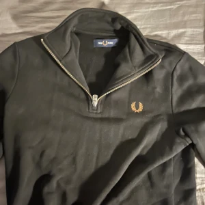 Svart half zip tröja från Fred Perry - Svart långärmad tröja från Fred Perry med silverfärgad half zip-dragkedja och broderad guldlogga på bröstet. Klassisk och stilren modell med ribbade muddar och hög krage. Perfekt för dig som gillar enkel men snygg streetstil. Köpt för 1300 
