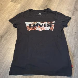 Levi's t-shirt - Svart t-shirt från Levi's med klassiskt logotryck framtill, där bakgrunden är fylld med blommigt mönster i rosa och beige toner. T-shirten har rund hals och korta ärmar. Perfekt för dig som gillar streetstyle och vill sticka ut lite extra. Knappt använd!