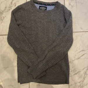 Grå kabelstickad tröja Bondelid - Kabelstickad grå tröja från Bondelid med rund halsringning och ribbade muddar. Klassisk och stilren modell som passar perfekt till jeans eller chinos. Mjuk och skön känsla, perfekt för kyligare dagar.