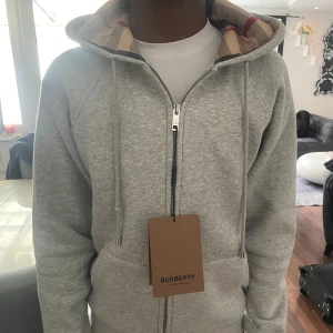 Grå hoodie från Burberry med dragkedja - Grå hoodie från Burberry med hel dragkedja och klassiskt rutigt foder i huvan. Tröjan har snörning i huvan, två fickor fram och diskret broderad logga på bröstet. Perfekt för en chill och stilren look.