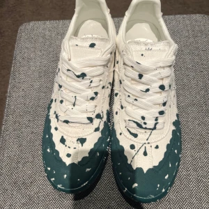 Maison Margiela sneakers med färgstänk - Maison Margiela sneakers i vitt tyg med gröna färgstänk och coola slitna kanter. Klassisk låg modell med snörning och vit sula. Unik design som sticker ut, perfekt för dig som gillar streetstyle och vill ha något annorlunda.