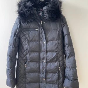 Svart pufferjacka från Vinson Polo Club - Säljer en svart pufferjacka från Vinson Polo Club med huva och fejkpäls. Jackan har quiltade detaljer på sidorna, dragkedjor på fickorna och en extra bröstficka med dragkedja. Perfekt för kalla dagar och riktigt snygg passform.