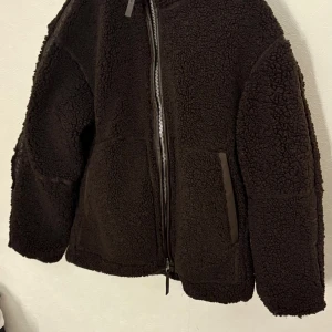  teddyjacka  - Mysig svart teddyjacka från H&M i storlek XS. Jackan har hög krage, två stora fickor framtill och stängs med dragkedja. Tillverkad i 100% återvunnen polyester och har en oversized passform som ger en cool och avslappnad vibe.