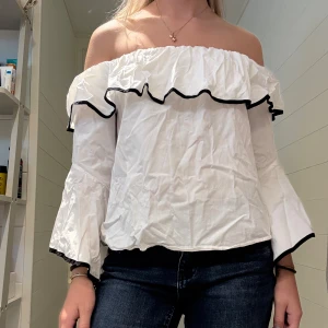 Vit offshoulder blus med volanger - Superfin vit offshoulder blus med stora volanger och svarta kanter. Aldrig använd. Storlek M men har stretch längs nacken/axlar. 