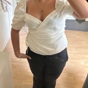 Vit omlottopp med puffärmar - Säljer en vit omlottopp med puffiga korta ärmar och knyt i sidan. Toppen har v-ringad urringning och är tillverkad i ett lätt bomullstyg. Perfekt för dig som gillar romantisk och trendig stil. Hör av dig vid frågor eller fler bilder!🥰🥰