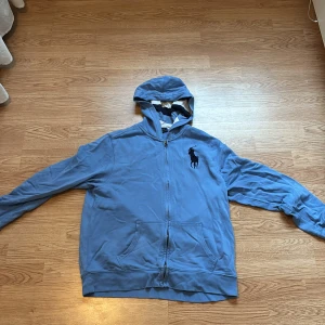 Blå hoodie från Polo Ralph Lauren XL - Blå hoodie från Polo Ralph Lauren i storlek XL med dragkedja och stor broderad logga på bröstet. Tröjan har huva med randigt foder och praktiska fickor framtill. Perfekt för en chill och avslappnad stil.