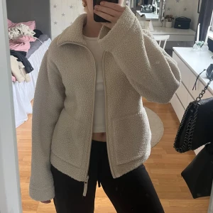 Beige teddyjacka med dragkedja - Mysig beige teddyjacka med hög krage och hel dragkedja framtill. Jackan har två stora fickor och är tillverkad i ett fluffigt fleece-material som håller dig varm. Perfekt för kyliga dagar och enkel att matcha med olika outfits.