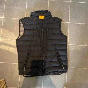 Svart dunväst från Parajumpers - Snygg svart dunväst från Parajumpers med quiltad design och hög krage. Västen har dragkedja framtill, två fickor med dragkedja och en cool patch på ryggen. Superlätt och perfekt för lager-på-lager-stil. Passar dig som vill ha en sportig och stilren look.