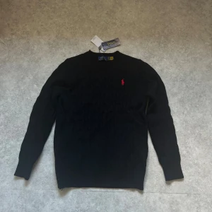 Svart stickad tröja från Polo Ralph Lauren - Svart stickad tröja från Polo Ralph Lauren med klassiskt kabelmönster och röd broderad logga på bröstet. Rund hals och långa ärmar. Perfekt för dig som gillar stilrena och tidlösa plagg med en touch av lyx.pris kan diskuteras