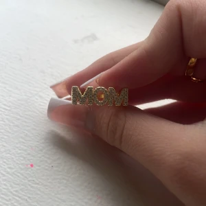 Guldig ring med texten MOM/WOW - Unik guldig ring med glittrande stenar som bildar texten 'MOM' från ena hållet och 'WOW' från andra. Ringen har en bred, fyrkantig form och är täckt av små gnistrande stenar för extra bling. Perfekt statement piece för dig som vill sticka ut. 