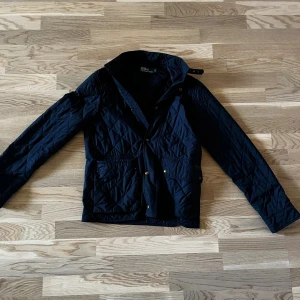 Quiltad jacka från Polo Ralph Lauren - Mörkblå quiltad jacka från Polo Ralph Lauren med klassisk krage, tryckknappar och två stora fickor framtill. Jackan har lång ärm och snygga detaljer som märkespatch och metallknappar. Perfekt för dig som gillar stilren och tidlös design. Använd en gång perfekt nu när de börjar bli lite kyligare 