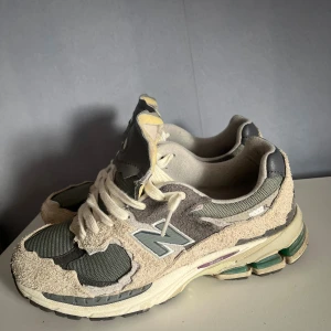 New Balance 2002R Protection Pack sneakers - Säljer ett par New Balance 2002R Protection Pack sneakers i grått och beige med coola slitna detaljer. Ovandelen är i mocka och mesh, med vita snören och gråa inslag. Skorna har en chunky sula med gröna och lila detaljer. Perfekta för dig som gillar streetwear vibes. Pris kan diskuteras vid rimliga förslag eftersom att dom bara är använda 4-5 ggr!