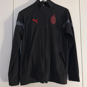 AC Milan träningsset från Puma svart - AC Milan träningsset från Puma i svart med röda detaljer. Jackan har hel dragkedja, ståkrage och långa ärmar med diskreta prickiga grå partier på axlarna. Byxorna har resår och dragsko i midjan samt röda loggor. Materialet är mjukt och sportigt, perfekt för fotbollsträning.