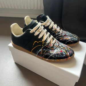 Maison Margiela sneakers paint splattern - Maison Margiela sneakers i svart skinn med unik färgstänkt design i vitt, rött, gult och blått. Klassisk låg modell med vita snören och beige gummisula. Insida i ljus skinn och märkning på sula och innersula. Perfekt för dig som vill sticka ut. Pris inte hugget i sten 