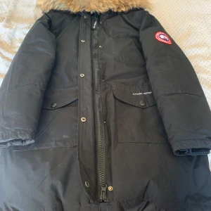 Svart Canada Goose parkas med päls - Svart parkas från Canada Goose med klassisk röd logotyp på ärmen och stor huva med päls. Jackan har flera fickor framtill, dragkedja och knappar. Perfekt för kalla vinterdagar och riktigt snygg streetstil.