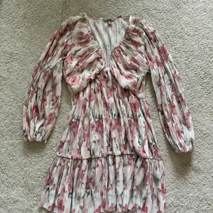 Blommig kortklänning med volang - Klänningen har v-ringad hals, långa ärmar med resår och volangdetaljer. Plisserad struktur ger en luftig och flowy känsla, perfekt för sommarens alla tillfällen. Storlek L