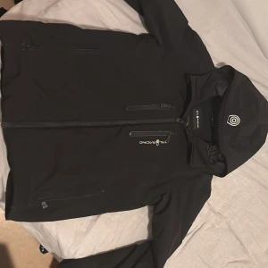 Svart vindjacka från Sail Racing - Svart vindjacka från Sail Racing med huva och dragkedja framtill. Jackan har två bröstfickor med dragkedja och diskret Sail Racing-logga på bröstet samt en broderad logga på huvan. Snygg och stilren modell som passar perfekt till blåsiga dagar.