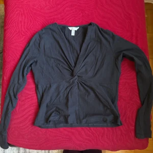 Svart omlottopp från H&M - Svart långärmad topp från H&M med snygg omlottdesign och twistad detalj framtill. Djup v-ringning ger en trendig look. Tillverkad i mjuk bomull som känns skön mot huden. Perfekt att styla med jeans eller kjol för en cool vibe.