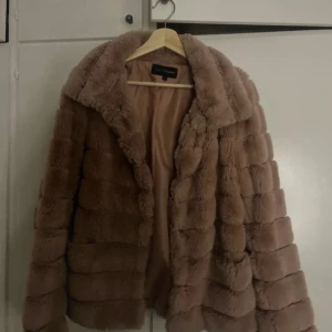 rosa beige pälsjacka från KappAhl - Supermjuk och fluffig beige pälsjacka från KappAhl. Jackan har bred krage, horisontella sömmar och är helfodrad för extra komfort. Perfekt för dig som vill ha en trendig och varm look under kyliga dagar.