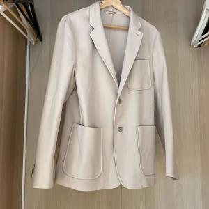 Beige kavaj med stora fickor - Stilren beige kavaj med två stora fickor framtill och en bröstficka. Klassisk krage och två knappar framtill. Perfekt för dig som gillar en clean och enkel look. Passar till många olika outfits.