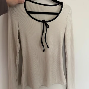 Beige ribbad långärmad topp med knyt - Säljer en beige ribbad långärmad topp med svart kant och knytband vid halsen. Toppen har en rund ringning och är i mjukt, stretchigt material. Perfekt för en stilren och enkel look.
