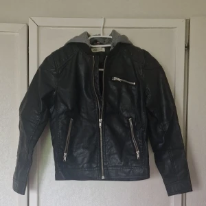 Svart skinnjacka med avtagbar huva från H&M - Svart fejkskinnjacka från H&M i storlek 140. Jackan har en grå huva som sitter fast med kardborre, flera dragkedjor framtill och på bröstet samt långa ärmar. Perfekt för dig som vill ha en edgy look med streetkänsla. Den är välanvänd jacka och slitage syns genom att tyget har rispats och på att kragen är nopprig.