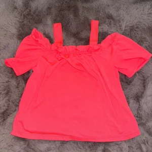 Neonrosa offshoulder topp - Snygg neonrosa topp med offshoulder-design och breda axelband. Toppen har korta ärmar och en lätt rynkad kant upptill som ger en cool och trendig look. Perfekt för dig som vill sticka ut med färg och stil.