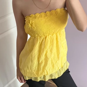 Gul bandeau tubtopp med blomdetalj - Söt gul bandeau tubtopp med stickad överdel och volangig, luftig nederdel. Toppen har en dekorativ blomma framtill och en vågig kant upptill. 