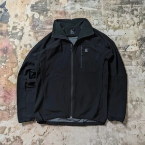 Svart vindjacka från Haglöfs M - Svart vindjacka från Haglöfs i sportig stil med hög krage och hel dragkedja framtill. Jackan har en bröstficka med dragkedja och diskret logga på både bröstet och ryggen. Materialet är lätt och känns smidigt, perfekt för uteliv och äventyr.