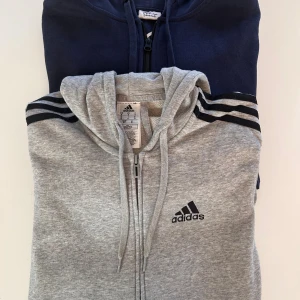 Adidas grå hoodie & blå Andy hoodie - Två hoodies säljes! En grå Adidas hoodie med svarta ränder på axlarna, dragkedja, snörning och huva. Den andra är en marinblå Andy by Pure Friday hoodie med dragkedja, huva och fickor. Båda är långärmade och i mjuk bomullsblandning.