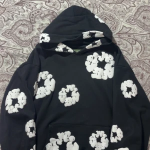Svart hoodie med vita blommönster - Svart hoodie med stora vita blommönster över hela tröjan, inklusive huvan. Tröjan har en klassisk huva och magficka framtill. Perfekt för dig som gillar streetwear och vill sticka ut med ett coolt mönster.