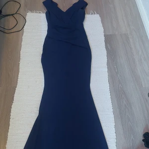 Marinblå långklänning från Bubbleroom  - Supersnygg marinblå långklänning från Goddiva (Bubbleroom) använd 1 gång, med v-ringad omlott-topp och breda axelband. Klänningen har figurnära passform och markerad midja, perfekt för bal eller fest. Materialet är mjukt och stretchigt för en bekväm känsla. Klänningen är i XS och jag är 160 o den släpar lite i golvet. Finns en liten fläck i klönningens ända (se 4), går nog att tvätta bort.