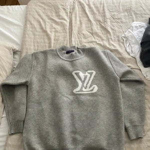 Grå stickad tröja från Louis Vuitton - Grå stickad tröja från Louis Vuitton med stort vitt LV-logo både fram och bak. Tröjan har rund halsringning, ribbade muddar och är långärmad. Perfekt för dig som vill ha en clean och lyxig look med streetvibe.