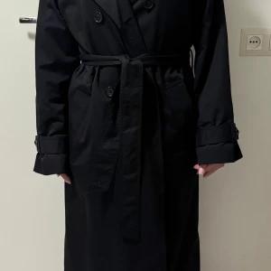 Svart lång trenchcoat med bälte - Stilren svart trenchcoat med klassisk dubbelknäppning, bälte i midjan och långa ärmar med spänndetaljer vid handlederna. Jackan har bred krage, två sidofickor och slits bak för extra rörelsefrihet. 