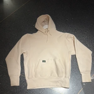 Beige hoodie från Camber x SNS - Säljer en beige hoodie från Camber for SNS, special edition. Tröjan har huva med snörning, känguruficka och ribbade muddar. Tillverkad i USA och gjord i mjuk bomull. Perfekt för dig som gillar clean och enkel streetwear. 