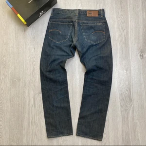 G-Star RAW blå jeans straight fit - Snygga blå jeans från G-Star RAW med klassisk femficksdesign och raka ben. Jeansen har kontrastsömmar, läderpatch med logga bak och diskret broderad detalj på bakfickorna. Tillverkade i slitstarkt denim med en mörk tvätt och subtila slitningar.
