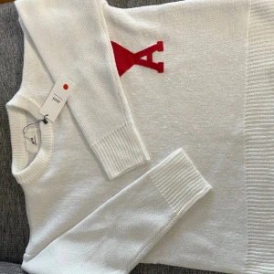 Vit stickad tröja från Ami - Vit stickad tröja från Ami med röd broderad logga i form av ett hjärta och bokstaven A på bröstet. Tröjan har rund halsringning, ribbade muddar och är långärmad. Perfekt för dig som vill ha en clean men ändå statement look.