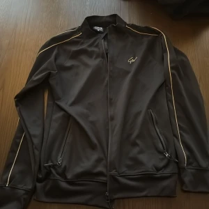 Svart track jacket från Gorilla Wear - Svart track jacket från Gorilla Wear med guldfärgade detaljer längs ärmarna och broderad GW-logga på bröstet. Jackan har dragkedja framtill, två fickor med dragkedja och en sportig, slim passform. Perfekt för dig som gillar streetwear och träning.
