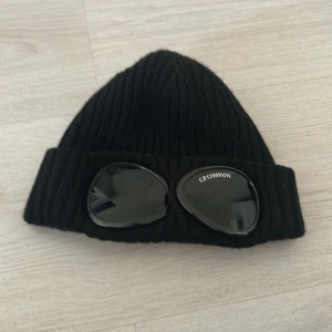 Svart Goggle-mössa från C.P. Company - Säljer en svart ribbstickad mössa från C.P. Company med de ikoniska goggle-linserna framtill. Mössan är tillverkad i mjuk ull och har en uppvikt kant där linserna sitter fast. Perfekt accessoar för dig som vill sticka ut.