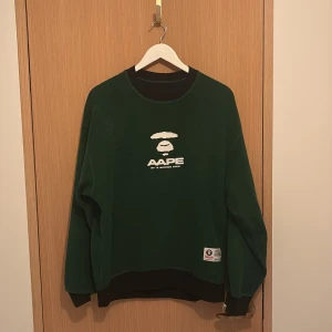 Grön sweatshirt från AAPE by A Bathing Ape - Mörkgrön sweatshirt från AAPE by A Bathing Ape med vit logga och text på bröstet. Svarta ribbade muddar vid hals, ärmslut och nederkant. Patch med AAPE-logga nertill. Perfekt för dig som gillar streetwear och vill sticka ut.