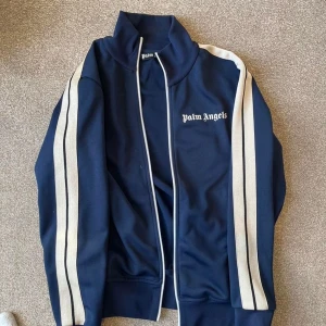 Blå track jacket från Palm Angels - Säljer en snygg marinblå track jacket från Palm Angels med vita ränder längs ärmarna och logga på bröstet. Jackan har hög krage, dragkedja framtill och sportig vibe. Perfekt för dig som gillar streetwear och vill sticka ut.