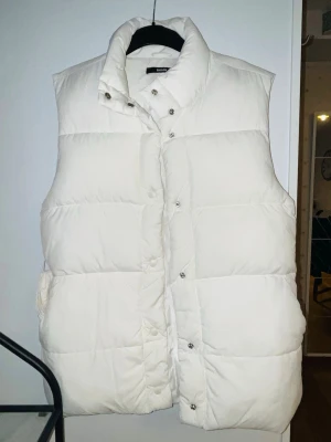Vit dunväst från Bik Bok - Säljer en fluffig och stilren vit dunväst från Bik Bok. Västen har djupa stora fickor, hög krage, och tryckknappar framtill. Perfekt att slänga över en hoodie eller tröja för extra värme.