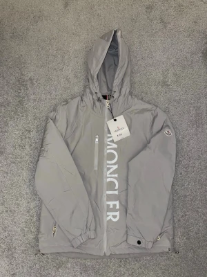 Grå vindjacka från Moncler med huva - Säljer en grå vindjacka från Moncler med stor logga framtill och klassisk patch på ärmen. Jackan har huva, dragkedja, bröstficka med zip och justerbara detaljer vid ärmslut och nederkant. Perfekt för dig som vill ha en clean och stilren look. Den är liten i storleken så den passar M 