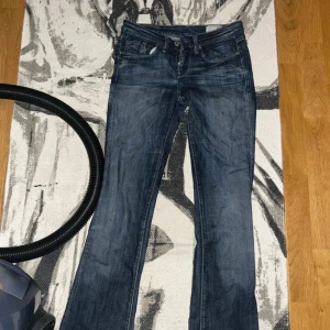 Blå bootcut jeans från G-Star RAW - Säljer ett par blå jeans från G-Star RAW med bootcut passform och råa, fransiga benslut. Klassisk femficksmodell med snygga slitningar och detaljerade sömmar. Jeansen har normal midja och är tillverkade i slitstarkt denimtyg.