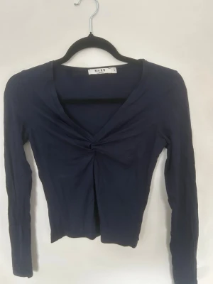 Mörkblå långärmad topp från NA-KD - Säljer en mörkblå långärmad topp från NA-KD i mjukt material. Toppen har en snygg v-ringning med twistad detalj framtill och är figurnära i modellen. Perfekt att styla med jeans eller kjol för en trendig look.
